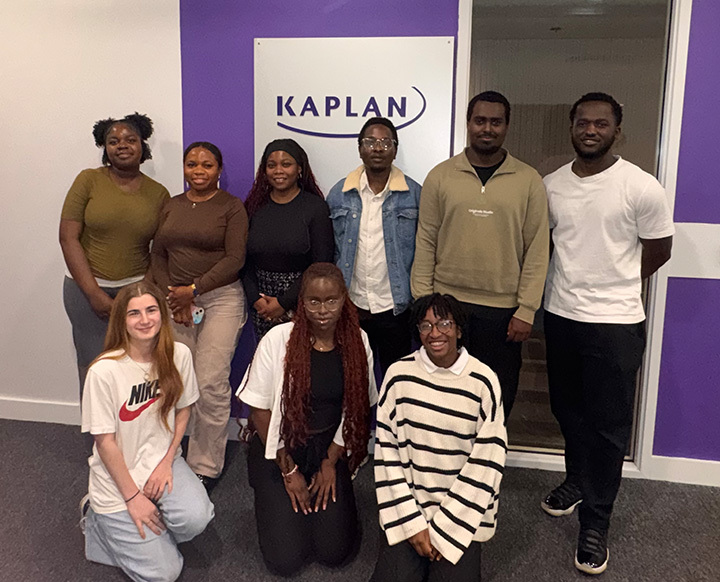 10KBI Interns A group of nine young adults posing together for a group photo in front of a Kaplan wall logo.