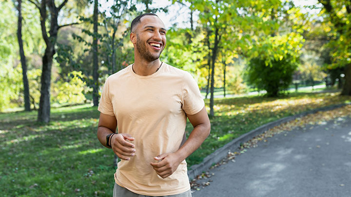 A smiling young man in a beige t-shirt jogging on a paved path in a sunlit park surrounded by green trees.