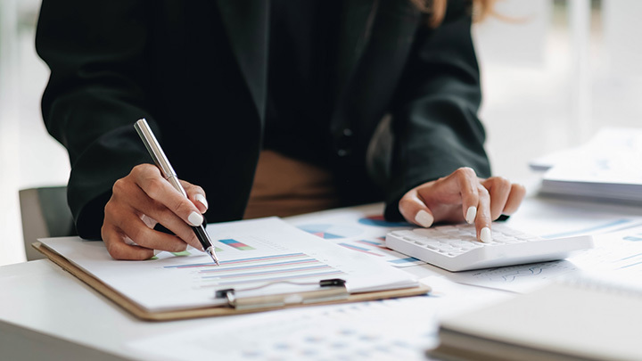 A person in a dark blazer reviewing financial charts on a clipboard while using a calculator, representing accountancy work and career development.