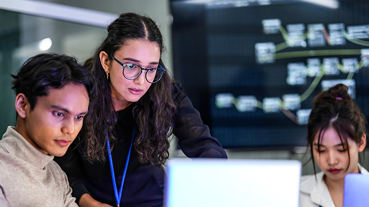 Three technology professionals collaborating around a laptop in a darkened office with data visualisation displayed on a large screen behind them, representing AI and data teamwork.