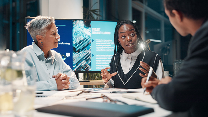 Three professionals in a meeting room having an engaged discussion with a machine learning presentation displayed on a screen behind them, representing AI strategy and business leadership.