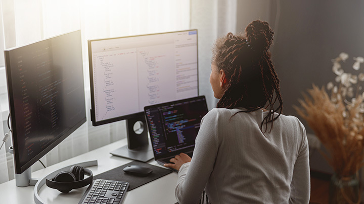 A woman with her back to the camera works at a desk with two large monitors displaying code, in a bright home office setting.