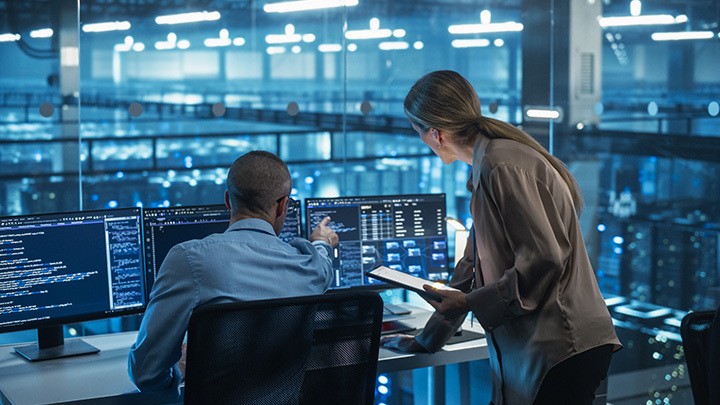 Two technology professionals working together at multiple screens displaying code in a large data centre environment, representing AI adoption and technical collaboration.