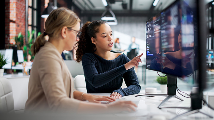 Two women collaborating at a computer screen in a modern office, representing AI upskilling and workforce development in a professional environment.