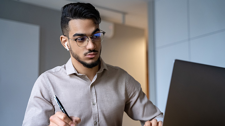 Man wearing glasses and earbuds looking at a laptop screen while taking notes.