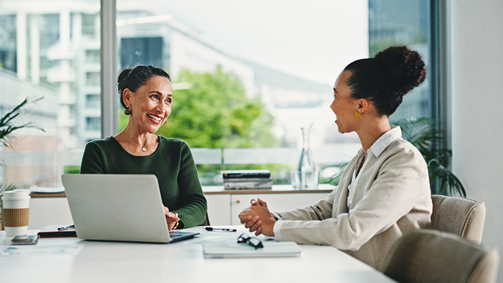 A line manager smiling and talking with an apprentice across a desk in a bright, modern office setting.