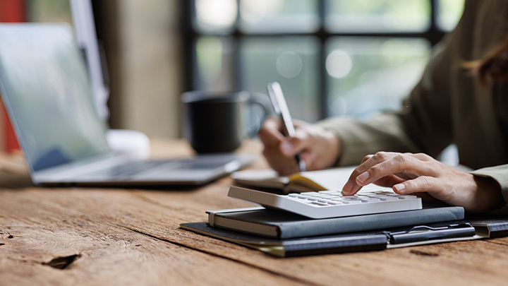 A person using a calculator and writing in a notebook next to a laptop on a wooden desk.