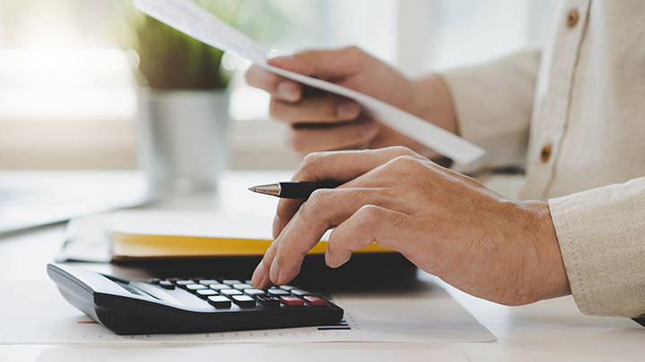 Close-up of a person's hands holding a financial document and using a desktop calculator at a brightly lit desk.