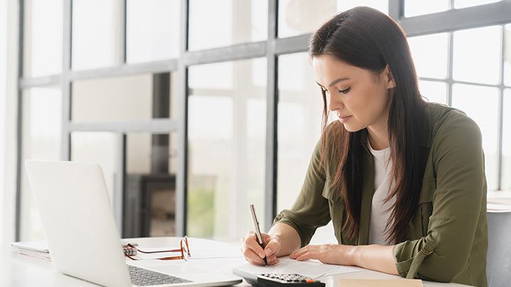 A woman with dark hair sitting at a bright desk, writing on paper with a laptop and calculator nearby.