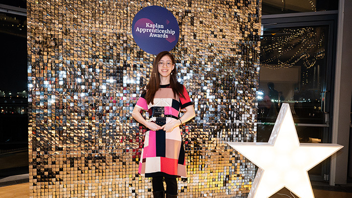 Chloe Auger holds her Apprentice of the Year trophy at the Kaplan Apprenticeship Awards ceremony, standing in front of a gold sequin backdrop displaying the Kaplan Apprenticeship Awards logo, with a large illuminated star to her right