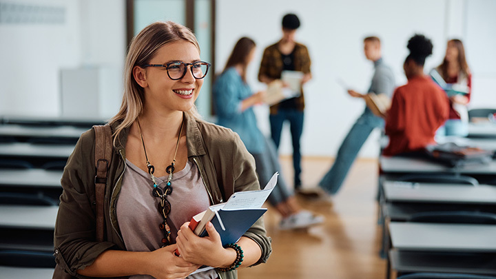 A smiling female student wearing glasses and holding notebooks in a classroom.