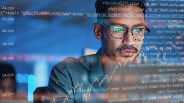A man wearing glasses focuses intently on multiple data charts and code displayed on a screen in front of him.
