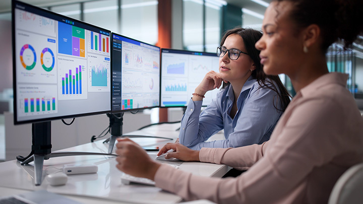 Two women collaborating at an office desk while looking at multiple computer screens displaying colorful data analytics dashboards and charts.