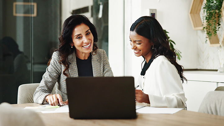 Two women smiling mid-discussion sat by a laptop in a work setting
