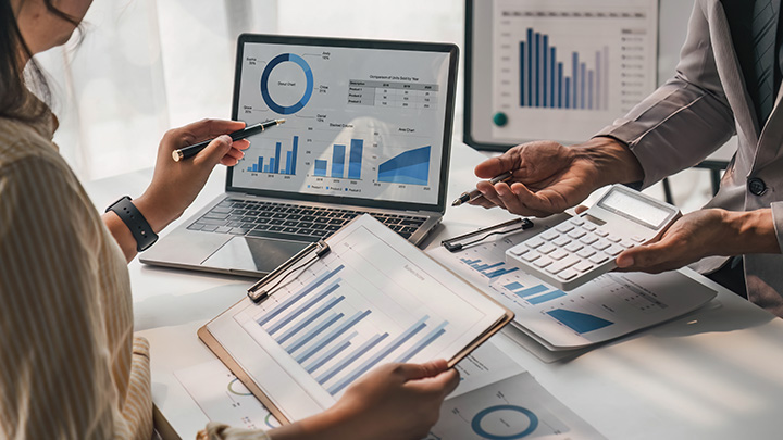 Two people review financial charts and graphs on a laptop, tablet, and printed reports while discussing figures and using a calculator.