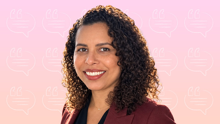 Headshot of a woman with curly brown hair wearing a dark red blazer against a light pink patterned background.