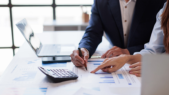 Two professionals in business attire collaborating over financial charts, data graphs, a calculator, and a laptop at a bright office desk.