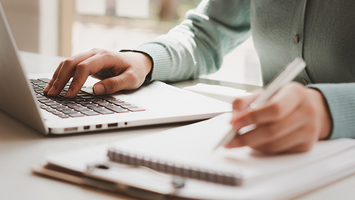 Hands working on a laptop and writing on a notepad with a pen