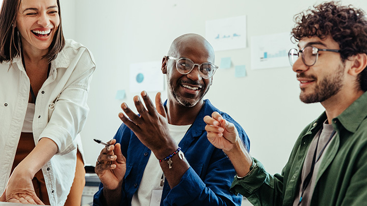 Three colleagues laughing and engaged in an active group discussion in a bright office environment.