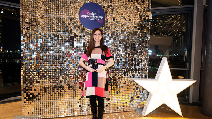 The winner of the Apprentice Of The Year award posing with the award at the ceremony, smiling in front of a sparkling backdrop with an event sign.