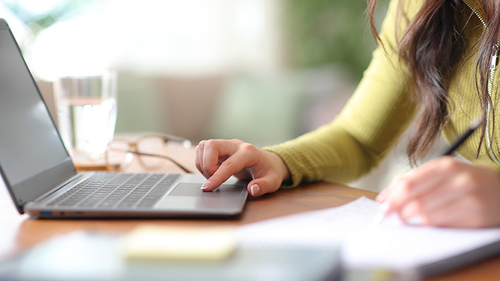 A person sitting at a wooden desk using a laptop touchpad with one hand while taking notes on paper with the other.
