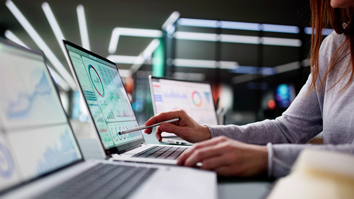 A woman with long red hair uses a stylus on a laptop displaying data dashboards and charts, with additional screens visible in a dark, modern office environment.