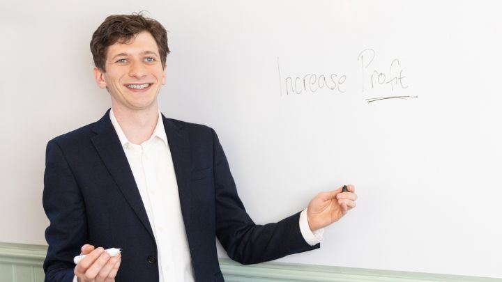 A man in a suit standing in front of a whiteboard with "Increase Profit" written on it, smiling and holding a marker.