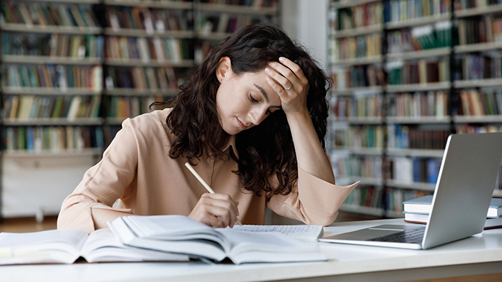 A woman sitting at a desk in a library, resting her head on her hand while studying, representing the pressures of balancing work and professional study.