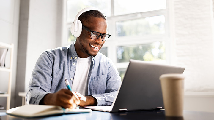 A young man wearing white headphones and glasses smiling as he works on a laptop.