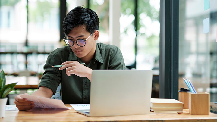 A young man wearing glasses reviews printed documents while working on a laptop at a wooden desk in a bright café-style setting.