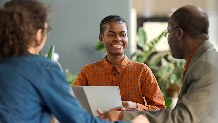 A young woman smiles while seated at a table with two colleagues during an office meeting.