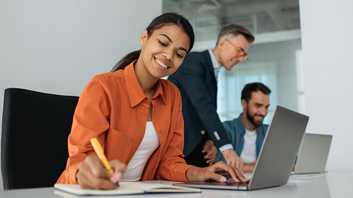 A smiling woman writes in a notebook while working on a laptop in an office, with two colleagues collaborating in the background.