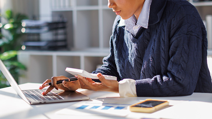 A person working at a desk, typing on a laptop while holding a pen and a white calculator, with financial documents and a smartphone nearby.