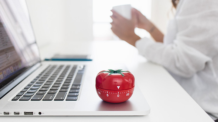 A red tomato-shaped kitchen timer sitting on an open laptop, with a person holding a mug in the blurred background.