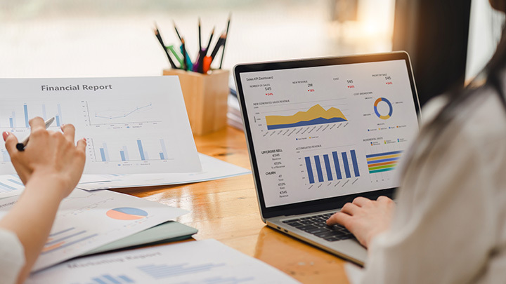 A person reviewing printed financial reports alongside an open laptop displaying colourful Power BI-style dashboards, including bar charts, line graphs, and KPI tiles, in a bright office setting.