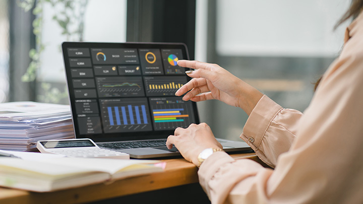 Woman pointing at a Power BI analytics dashboard on a laptop, surrounded by paperwork and a calculator on a wooden desk.