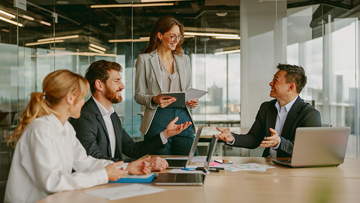 A group of four professionals in a meeting room, with a woman standing and presenting to engaged colleagues around a table, representing workplace communication and collaboration.