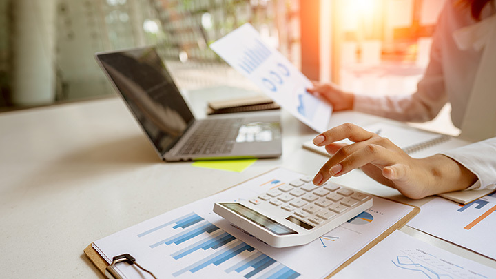 Woman's hand over a calculator, on a clipboard with bar charts on it, next to a laptop