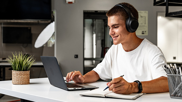 A young man wearing headphones sitting at a white desk, smiling while working on a laptop and taking notes in a notebook.