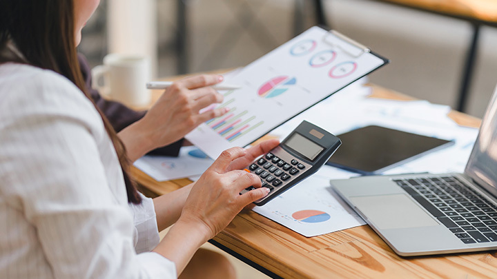 A woman reviewing financial charts and graphs while using a calculator at a desk, representing business analysis and problem-solving in a professional role.