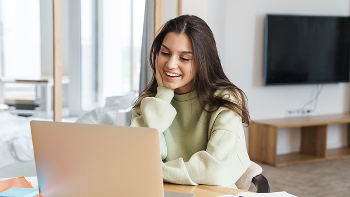 A smiling young woman wearing a light green sweater sitting at a desk and looking at her laptop screen.