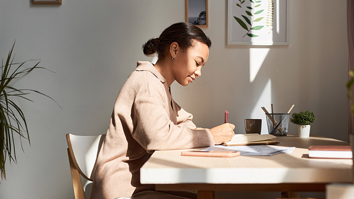 A young woman sitting at a desk in a sunlit room, writing in a notebook.