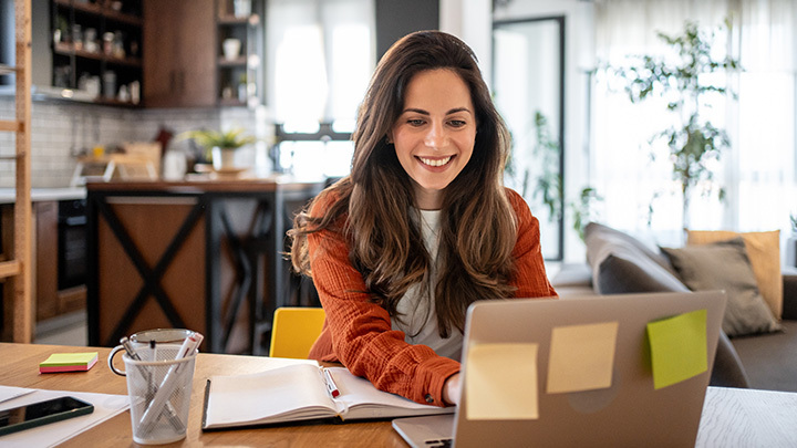 A smiling woman sitting at a table working on a laptop at home.