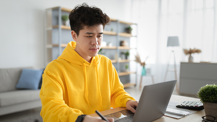 A young man wearing a bright yellow hoodie sitting at a desk, looking focused while working on a laptop and holding a pen.