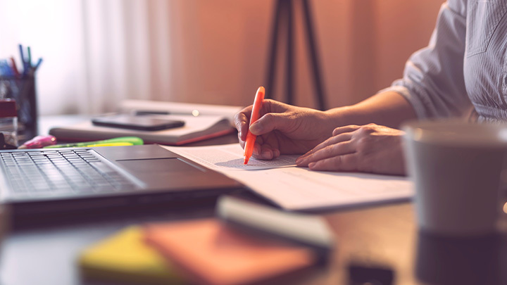 A person writes notes with an orange pen at a desk beside an open laptop, with colourful stationery and notebooks visible in a warm, softly lit setting.