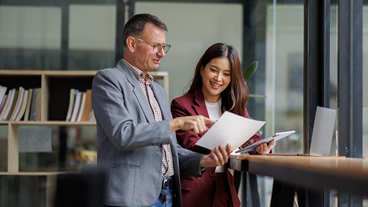 Two professionals standing in a modern office, reviewing a document together while discussing work.