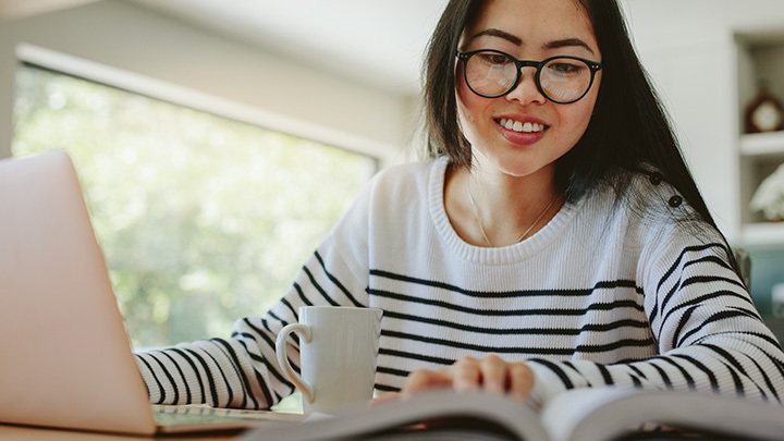 A smiling woman with round black glasses sitting at a table, using a laptop with an open book in front of her.