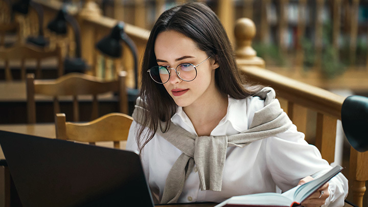 A young woman studying at a laptop while holding an open book.
