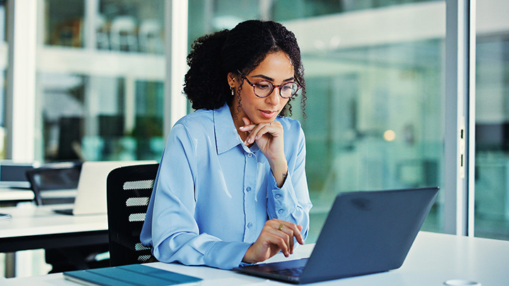 A woman wearing glasses and a blue button-down shirt working thoughtfully on a laptop at a bright office desk.