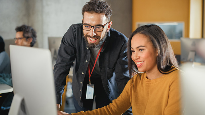 A male mentor in glasses leans over to guide a smiling female colleague at a desktop computer in a modern office, with a colleague visible in the background.